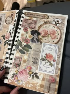 a hand holding an open journal with flowers and pictures on the pages, in front of a clock