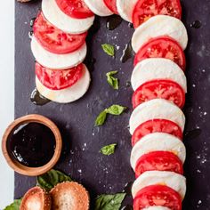 sliced tomatoes, mozzarella and basil on a black cutting board with bread in the background