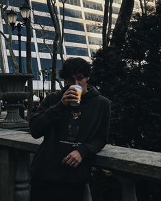 a young man drinking from a cup while standing next to a park bench in front of tall buildings