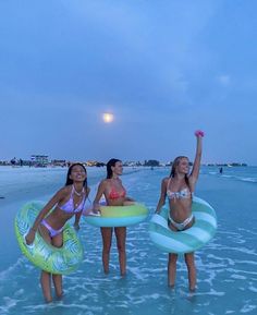 three women in bikinis are standing on the beach with boogie boards and float rings