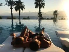 two women laying on the edge of a swimming pool with palm trees in the background