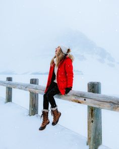 a woman sitting on a fence in the snow wearing boots and a red parka