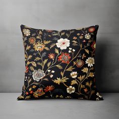 a black pillow with colorful flowers and leaves on the front, sitting against a gray wall