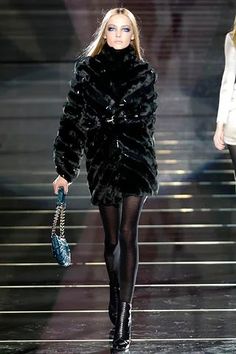 a model walks down the runway in a black fur coat and tights with high heels