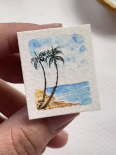 a hand holding a small piece of paper with a painting of a palm tree on it