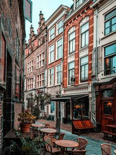 tables and chairs are lined up on the sidewalk in front of old brick buildings with potted plants