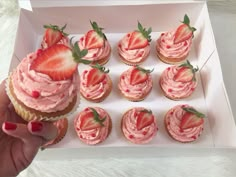 a person is holding up a box of cupcakes with pink frosting and strawberries on top