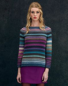 a woman standing in front of a black wall wearing a multicolored sweater and purple skirt