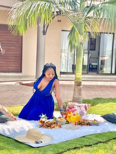 a woman in a blue dress is sitting on a blanket with food and drinks near a palm tree