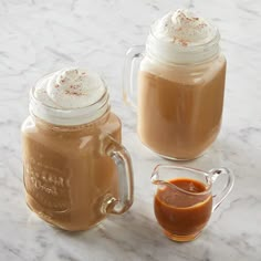 two mason jars filled with hot chocolate and topped with whipped cream, sit on a marble countertop