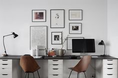 a white desk topped with a computer monitor and two wooden chairs next to a wall filled with pictures