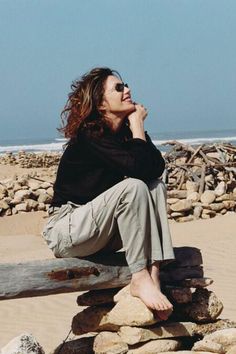 a woman sitting on top of a pile of rocks next to the ocean with her hand under her chin