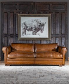 a brown leather couch sitting in front of a wooden paneled wall with a buffalo drawing on it