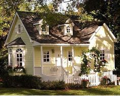 a small yellow house with white trim on the roof and windows, surrounded by greenery