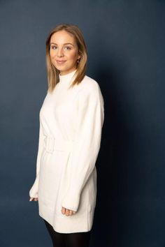 a woman standing in front of a blue wall wearing a white sweater and black leggings