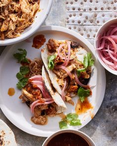 two white plates topped with tacos next to bowls of sauce and onions on a table