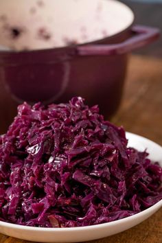 a white bowl filled with red cabbage on top of a wooden table next to a purple casserole dish