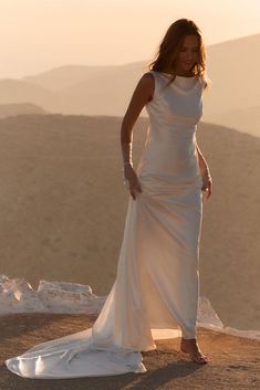 a woman in a long white dress standing on top of a hill with her hands behind her back