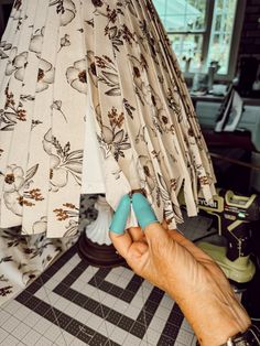 a hand holding an object in front of a curtain with flowers on it and a window behind it