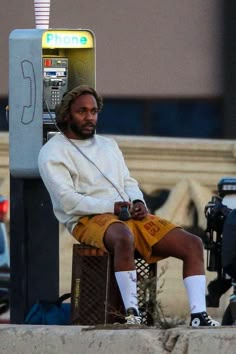 a man sitting on top of a parking meter next to a phone booth with his hands in his pockets