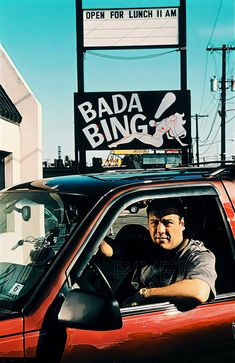 a man sitting in the driver's seat of a red car next to a bada bing sign