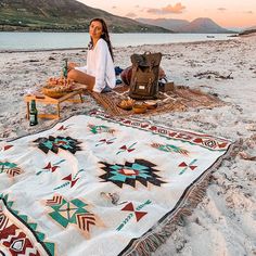 a woman sitting on the beach next to a picnic blanket with food and drinks in front of her