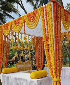 an outdoor wedding setup with yellow and orange flowers on the canopy, white tablecloths and pillows