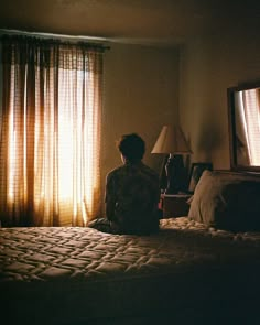 a woman sitting on top of a bed next to a window covered in sheer curtains