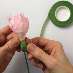 two hands are holding a pink flower next to a roll of tape and a green spool