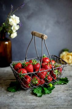 a basket filled with lots of ripe strawberries on top of a wooden table next to flowers