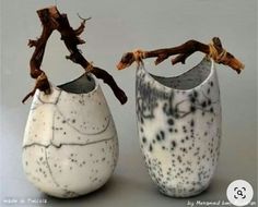 two white vases with branches in them on a gray surface, one has black spots and the other is speckled