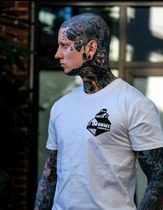 a man with tattoos on his face and neck standing in front of a building wearing a white t - shirt