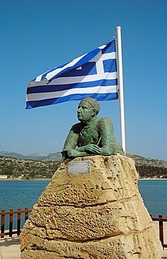 a statue with a greek flag flying in the wind next to a body of water