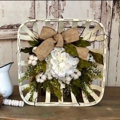 a white flower arrangement in a basket with burlocks and greenery on a table