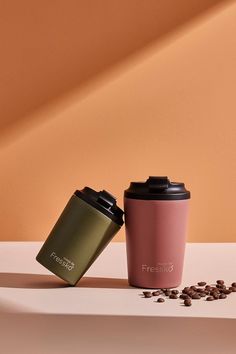 two coffee cups sitting next to each other on top of a white table with coffee beans scattered around them