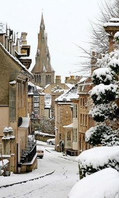 a snowy street with buildings and trees in the foreground, and a church steeple in the background