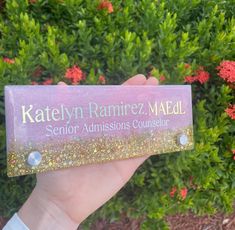 someone holding up a pink and gold business card in front of some bushes with red flowers