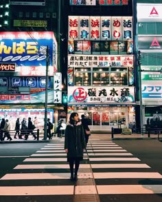 a woman walking across a cross walk in the middle of a city at night time