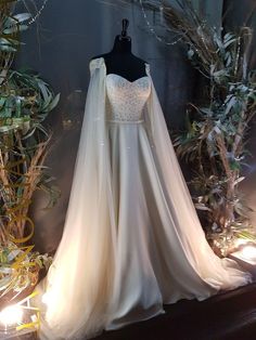 a white wedding dress on display in front of some plants and branches with lights coming from behind it