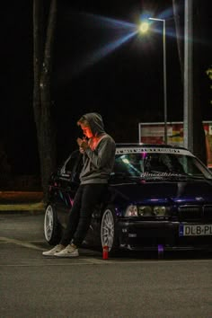 a man sitting on the hood of a car talking on his cell phone at night