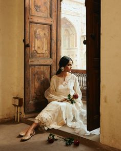 a woman sitting on a bench in front of a wooden door wearing a white dress