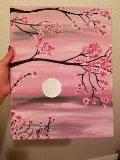 a person is holding up a painting with pink flowers and a full moon in the background