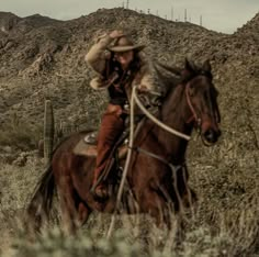 a man riding on the back of a brown horse next to a cactus filled hillside