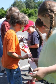 several children are standing in a parking lot and one girl is handing something out to the other