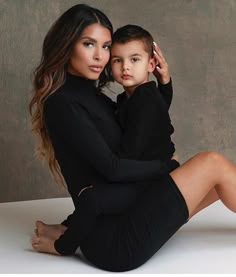 a woman sitting next to a little boy on top of a white table in front of a gray wall