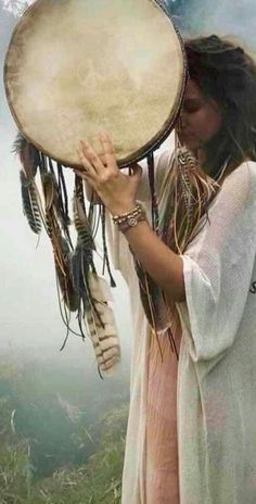 a woman with long hair holding a large drum in front of her face while standing on top of a hill