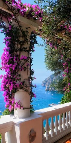 purple flowers are growing on the side of a white building near water and mountains in the background