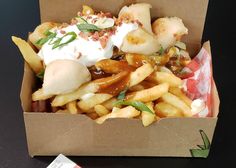 a cardboard box filled with french fries covered in sauce and toppings, sitting on a table