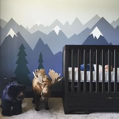 two baby cribs with mountains painted on the wall behind them and a black cat standing next to one