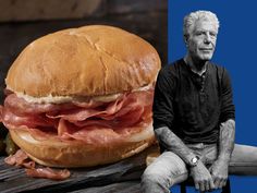 an older man sitting on a bench next to a large sandwich with meat and cheese